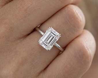 May include: A silver-toned ring featuring a large, rectangular emerald-cut clear gemstone set in a four-prong setting. The band is thin and polished, showcasing a clean, reflective surface that highlights the faceted stone's geometric clarity.