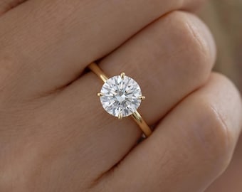May include: A solitaire ring featuring a round brilliant-cut clear gemstone set in a polished yellow gold band. The stone is secured by four thin prongs, showcasing a faceted surface that reflects light with a bright, white sparkle.