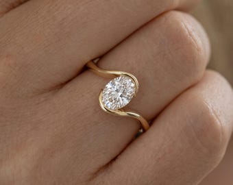 May include: A gold ring featuring a single oval-cut clear gemstone set in a bypass style band. The metal band curves around the top and bottom of the central stone, creating a sleek, continuous design that highlights the faceted white stone.
