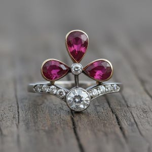 May include: A two-piece ring set featuring a floral design. The top ring has three teardrop-shaped, magenta gemstones with gold settings, and a small diamond. The bottom ring has a large, round diamond and smaller diamonds.