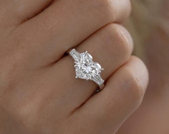 May include: A silver-toned ring featuring a large, central heart-shaped gemstone flanked by two smaller, elongated baguette-cut stones on either side, all set on a polished metal band.