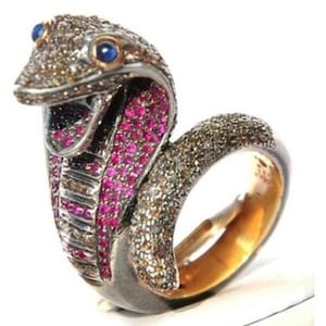 May include: A cobra-shaped ring with a silver and gold band. The snake's head is adorned with small, sparkling stones and blue eyes. The cobra's mouth is lined with pink gemstones.