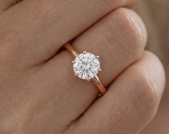May include: A solitaire ring featuring a round brilliant-cut clear gemstone set in a polished rose gold band. The stone is secured by six prongs, creating a classic profile. The metal band has a smooth, rounded finish and a warm pinkish hue.