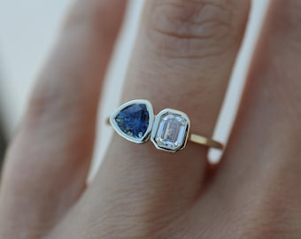 May include: A gold ring featuring two gemstones: a triangular blue sapphire and a rectangular, clear emerald-cut diamond. The gemstones are set side-by-side in a bezel setting. The ring is shown on a person's finger.