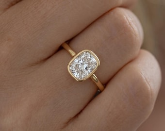 May include: A gold ring featuring a cushion-cut clear gemstone set in a smooth bezel setting. The band is thin and polished, crafted from yellow gold, and the center stone displays multiple facets that reflect light from various angles.