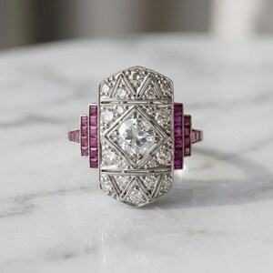 May include: An Art Deco style ring with a central round diamond and smaller diamonds set in a geometric pattern. The ring features rectangular ruby accents on each side, set in a silver or white gold band. The ring is displayed on a marble surface.