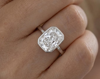 May include: A silver-toned ring featuring a large, rectangular radiant-cut clear gemstone set in a smooth bezel setting. The stone has multiple facets that reflect light, and the band is a simple, polished metal finish that encircles the finger.
