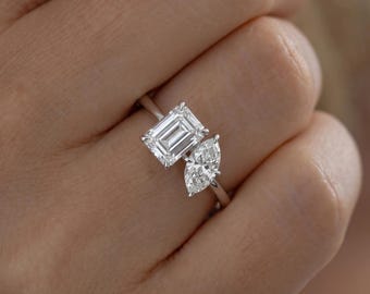 May include: A silver-toned ring featuring two prominent clear stones set on a thin band. One stone is rectangular with emerald-cut facets, while the adjacent stone is marquise-shaped with a pointed end, both secured by metal prongs.