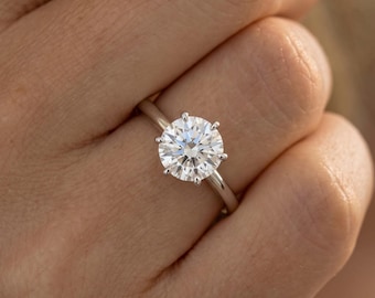May include: A solitaire ring featuring a round brilliant-cut clear gemstone set in a polished silver-toned metal band. The stone is secured by six prongs, creating a classic circular profile that reflects light across its faceted surface.