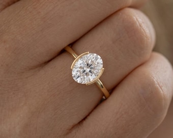 May include: A gold ring featuring a large, oval-cut, clear white gemstone set in a partial bezel setting. The band is thin and polished, crafted from yellow gold, and the stone is positioned horizontally across the finger with open sides.