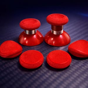 May include: A set of red gaming controller thumbsticks and replacement pads. The thumbsticks have a textured top and a silver band. The pads are round and triangular, all with a textured surface. The items are on a dark blue surface.