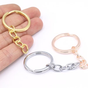 Puede incluir: Llaveros en tonos oro, plata y oro rosa. Cada llavero presenta un anillo circular conectado a una cadena. Un llavero dorado es sostenido por una mano. Los llaveros están sobre un fondo blanco.