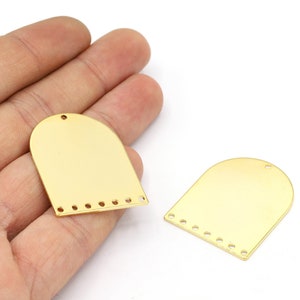 Könnte beinhalten: Zwei goldfarbene, bogenförmige Rohlinge aus Metall mit Löchern am unteren Rand. Die Rohlinge liegen auf einer Hand.