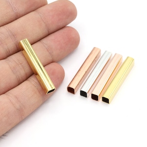Pode incluir: Uma coleção de tubos metálicos retangulares em várias cores, incluindo dourado, cobre, prateado e ouro rosa. Um tubo dourado é segurado em uma mão, enquanto os outros estão dispostos em uma superfície branca. Estes tubos são provavelmente componentes de joias.