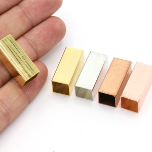 Pode incluir: Cinco tubos quadrados de metal em diferentes cores: ouro, prata, cobre, ouro rosa e latão. Os tubos são ocos e têm um acabamento liso.