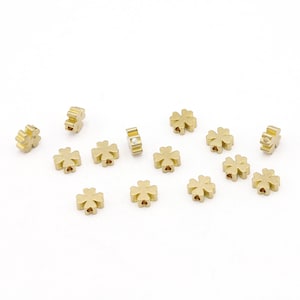 May include: A collection of 13 gold-colored four-leaf clover charms. The charms are small and have a slightly raised design. One charm has a small white stone in the center.