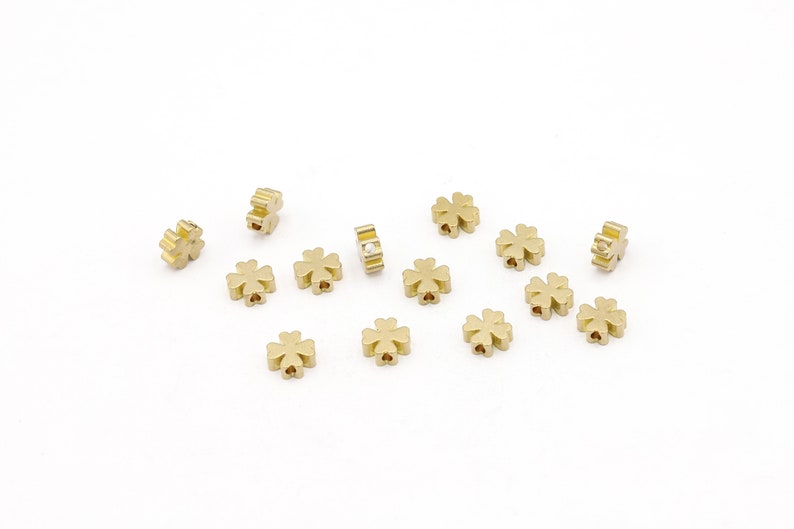 May include: A collection of 13 gold-colored four-leaf clover charms. The charms are small and have a slightly raised design. One charm has a small white stone in the center.