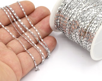 Könnte beinhalten: Eine Spule mit silberfarbener Kette mit rechteckigem Glieddesign. Die Kette ist teilweise abgewickelt, wobei ein Teil in einer Hand gehalten wird. Die Kette befindet sich auf einer weißen Spule.