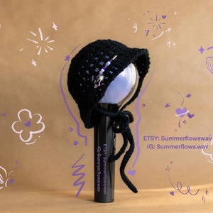 May include: Black crochet hat with a decorative pin, tied with strings. The hat is placed on a black cylinder. The background is tan with white and purple doodles. Etsy and Instagram handles are visible.