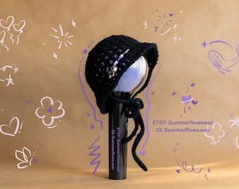 May include: Black crochet hat with a decorative pin, tied with strings. The hat is placed on a black cylinder. The background is tan with white and purple doodles. Etsy and Instagram handles are visible.
