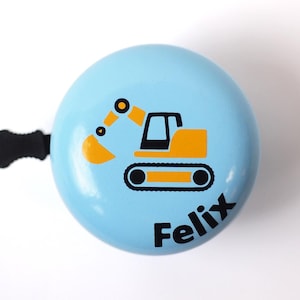 Peut inclure: Une sonnette de v&eacute;lo bleu clair avec un dessin d'excavatrice noir et jaune et le nom "Felix" imprim&eacute; sur le devant.