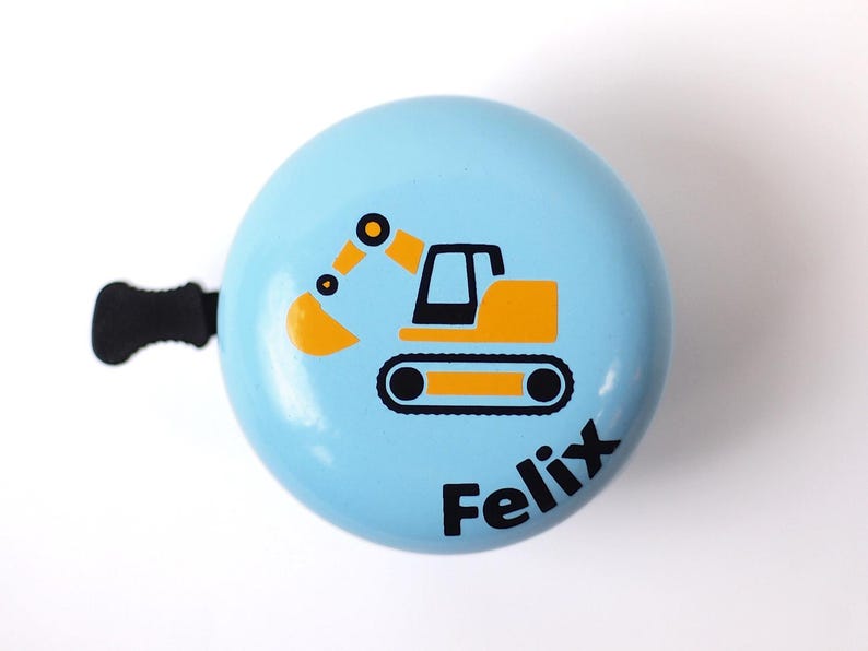Peut inclure: Une sonnette de v&eacute;lo bleu clair avec un dessin d'excavatrice noir et jaune et le nom "Felix" imprim&eacute; sur le devant.