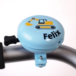 Peut inclure: Une sonnette de v&eacute;lo bleue avec un dessin d'excavatrice jaune et le nom "Felix" imprim&eacute; dessus.