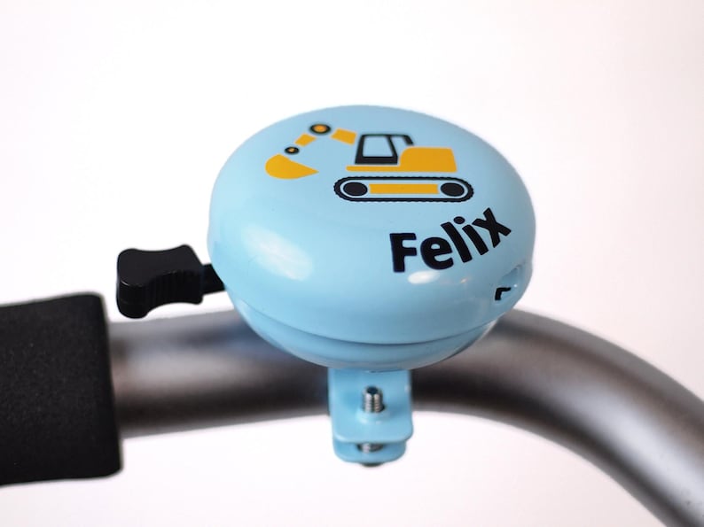 Peut inclure: Une sonnette de v&eacute;lo bleue avec un dessin d'excavatrice jaune et le nom "Felix" imprim&eacute; dessus.