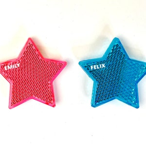 Könnte beinhalten: Zwei sternförmige reflektierende Sicherheitsaufkleber, einer rosa mit dem Namen "EMILY" und einer blau mit dem Namen "FELIX".