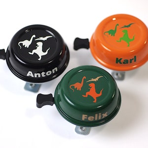 Könnte beinhalten: Drei Fahrradklingeln mit Dinosaurier-Motiven. Die Klingeln sind schwarz, orange und grün. Die Klingeln sind mit den Namen Anton, Karl und Felix personalisiert.