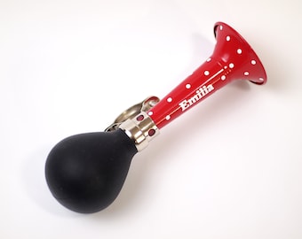 Peut inclure: Un klaxon de vélo rouge à pois blancs avec le nom "Emilia" inscrit dessus. Le klaxon a une poire en caoutchouc noire.