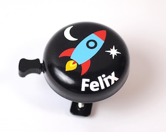 Peut inclure: Sonnette de vélo noire avec un motif de fusée. La sonnette présente une fusée bleue avec des ailerons rouges et une flamme jaune, un croissant de lune blanc et une étoile. Le nom "Felix" est imprimé en blanc.