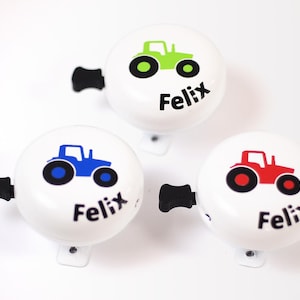 Könnte beinhalten: Drei weiße Fahrradklingeln mit Traktor-Design und dem Namen "Felix" auf jeder. Die Traktoren sind grün, blau und rot.