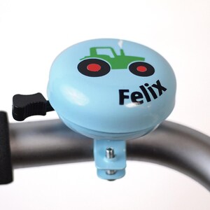 Peut inclure: Une sonnette de v&eacute;lo bleu clair avec un dessin de tracteur vert et le nom "Felix" imprim&eacute; dessus.
