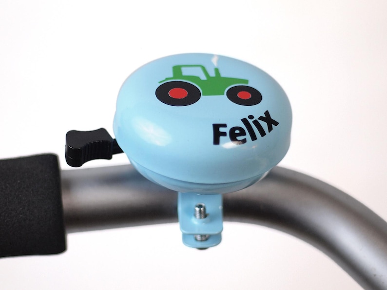 Peut inclure: Une sonnette de v&eacute;lo bleu clair avec un dessin de tracteur vert et le nom "Felix" imprim&eacute; dessus.