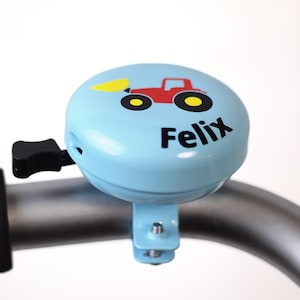 Peut inclure: Une sonnette de v&eacute;lo bleu clair avec un graphique de tracteur rouge et le nom "Felix" imprim&eacute; dessus.
