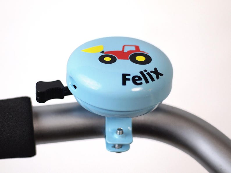 Peut inclure: Une sonnette de v&eacute;lo bleu clair avec un graphique de tracteur rouge et le nom "Felix" imprim&eacute; dessus.