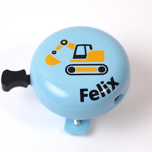 Peut inclure: Une sonnette de v&eacute;lo bleue avec un graphique d'excavatrice jaune et le nom "Felix" imprim&eacute; en noir.