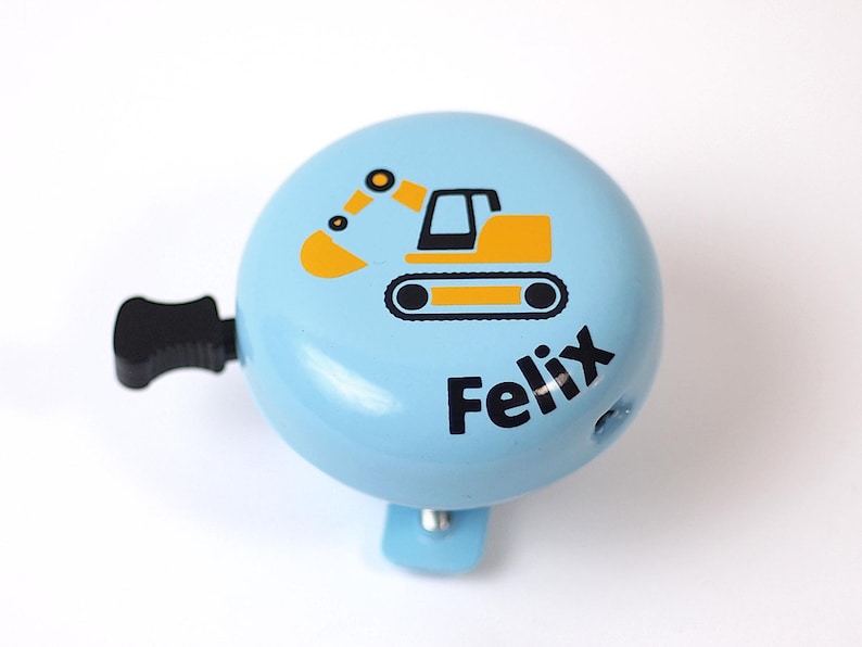 Peut inclure: Une sonnette de v&eacute;lo bleue avec un graphique d'excavatrice jaune et le nom "Felix" imprim&eacute; en noir.