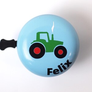 Peut inclure: Une sonnette de v&eacute;lo bleu clair avec un dessin de tracteur vert et le nom "Felix" imprim&eacute; sur le devant.