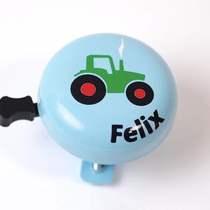 Peut inclure: Une sonnette de v&eacute;lo bleu clair avec un dessin de tracteur vert et le nom "Felix" imprim&eacute; sur le devant.