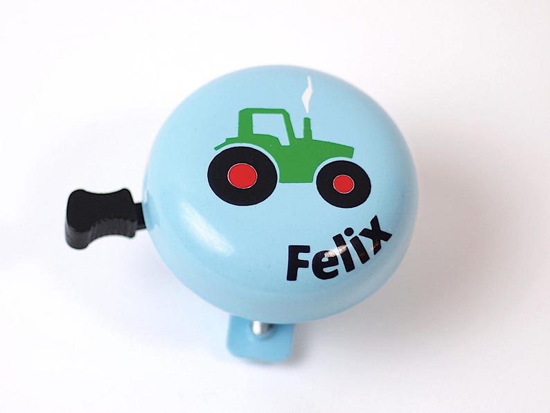 Peut inclure: Une sonnette de v&eacute;lo bleu clair avec un dessin de tracteur vert et le nom "Felix" imprim&eacute; sur le devant.