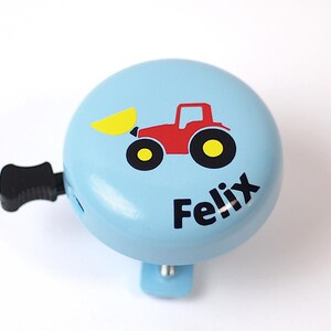 Peut inclure: Une sonnette de v&eacute;lo bleu clair avec un tracteur rouge et une pelle jaune. La sonnette a une poign&eacute;e noire et le nom "Felix" est imprim&eacute; sur le devant.