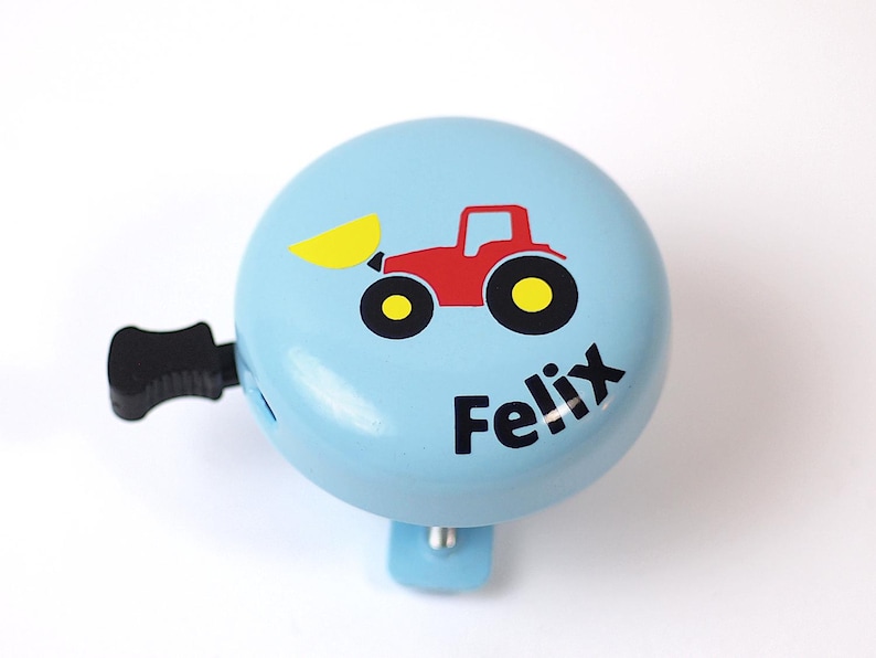 Peut inclure: Une sonnette de v&eacute;lo bleu clair avec un tracteur rouge et une pelle jaune. La sonnette a une poign&eacute;e noire et le nom "Felix" est imprim&eacute; sur le devant.