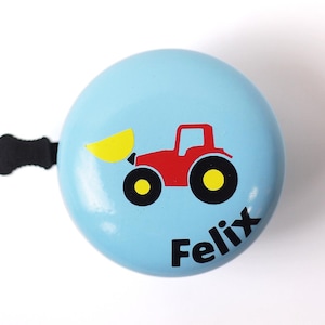 Peut inclure: Une sonnette de v&eacute;lo bleu clair avec un tracteur rouge et une pelle jaune. La sonnette porte le nom "Felix" imprim&eacute; dessus.
