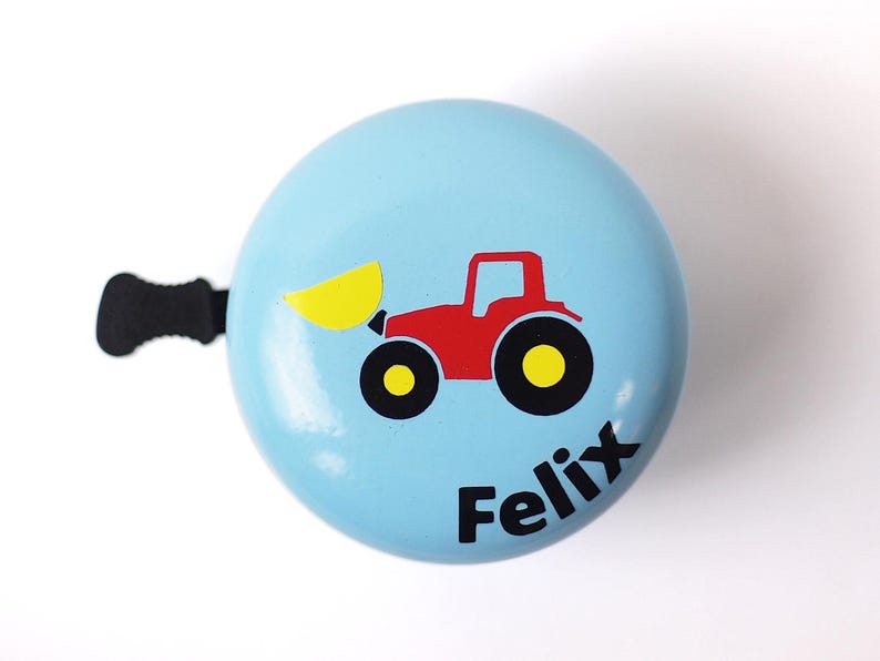 Peut inclure: Une sonnette de v&eacute;lo bleu clair avec un tracteur rouge et une pelle jaune. La sonnette porte le nom "Felix" imprim&eacute; dessus.