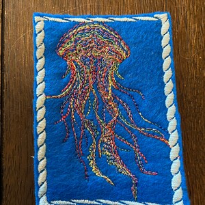 May include: A colorful embroidered jellyfish on a blue felt background with a white rope border.