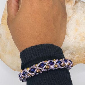 Puede incluir: Una pulsera de cuentas con un patrón de celosía entrecruzado de pequeñas cuentas de color violeta claro iridiscente, intercaladas con cuentas esféricas más grandes en oro metálico y azul real profundo, creando un accesorio texturizado y colorido.