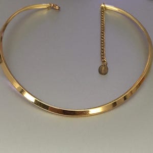 Puede incluir: Un collar tipo gargantilla de metal en tono dorado con un diseño de banda curva, lisa y plana. El collar incluye una extensión de cadena corta y delicada en un extremo, terminada con una pequeña etiqueta metálica circular colgante.