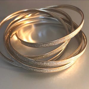 May include: A collection of gold and silver bangles, intertwined and resting on a light-colored surface. Some bangles have a glittery texture, reflecting light. The jewelry is arranged in a circular pattern, showcasing the design.